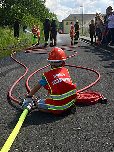 2019 - 35. Leistungsnachweis der Feuerwehren des Rheinisch-Bergischen Kreis Ein Kind in Feuerwehrkleidung sitzt auf der Straße und hält einen Wasserschlauch. Im Hintergrund sind weitere Kinder in Feuerwehruniformen und Erwachsene zu sehen. Die Szene spielt sich an einem sonnigen Tag ab, mit Wasser, das aus Schläuchen sprüht.