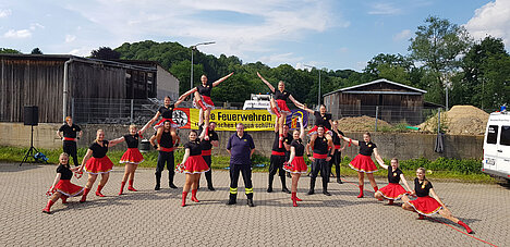2019 - 35. Leistungsnachweis der Feuerwehren des Rheinisch-Bergischen Kreis Eine Gruppe von Tänzern in roten Röcken und schwarzen Oberteilen posiert in verschiedenen Formationen in einer Außenumgebung. Im Hintergrund sind einige Gebäude und eine Feuerwehr-Flagge sichtbar. Die Gruppe zeigt eine lebhafte Darbietung.