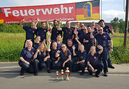 2019 - 35. Leistungsnachweis der Feuerwehren des Rheinisch-Bergischen Kreis Eine Gruppe von Feuerwehrmitgliedern posiert lächelnd vor einem großen Banner mit der Aufschrift „Feuerwehr“. Sie feiern vermutlich einen Erfolg, da sie Pokale in der Mitte halten. Die Mitglieder tragen einheitliche schwarze T-Shirts und zeigen ihre Freude mit hochgehobenen Händen.