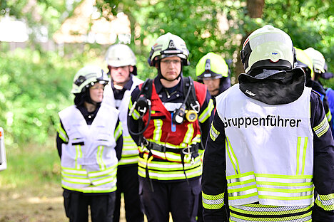 2023 - 38. Leistungsnachweis der Feuerwehren des Rheinisch-Bergischen Kreis Eine Gruppe von Einsatzkräften in Feuerwehruniformen versammelt sich im Freien. Einige tragen Helme und reflektierende Westen. Ein Gruppenführer steht im Vordergrund, auf dessen Weste "Gruppenführer" steht. Die Umgebung ist bewaldet.