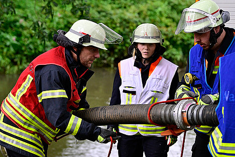 2023 - 38. Leistungsnachweis der Feuerwehren des Rheinisch-Bergischen Kreis Drei Feuerwehrleute arbeiten zusammen, um einen Schlauch an eine Wasserpumpe anzuschließen. Sie tragen Schutzkleidung und Helme, während sie sich auf die Aufgabe konzentrieren. Im Hintergrund ist eine grüne Umgebung und Wasser sichtbar.