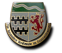Ehrennadel in Gold für besondere Verdienste im Feuerwehrwesen Das Bild zeigt ein Wappen mit grüner und grauer Farbgebung. Auf der linken Seite befinden sich schwarze Wellenlinien und auf der rechten Seite ein roter Löwe. Unten steht der Schriftzug „Für besondere Verdienste im Feuerwehrwesen“.
