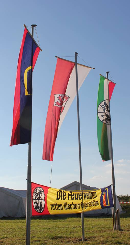Drei Fahnen auf Fahnenmasten stehen vor einem klaren Himmel. Die Fahnen repräsentieren verschiedene Wappen oder Symbole, während ein großes Banner mit der Aufschrift „Die Feuerwehr retten – löschen – schützen“ in gelber Farbe am Boden hängt.