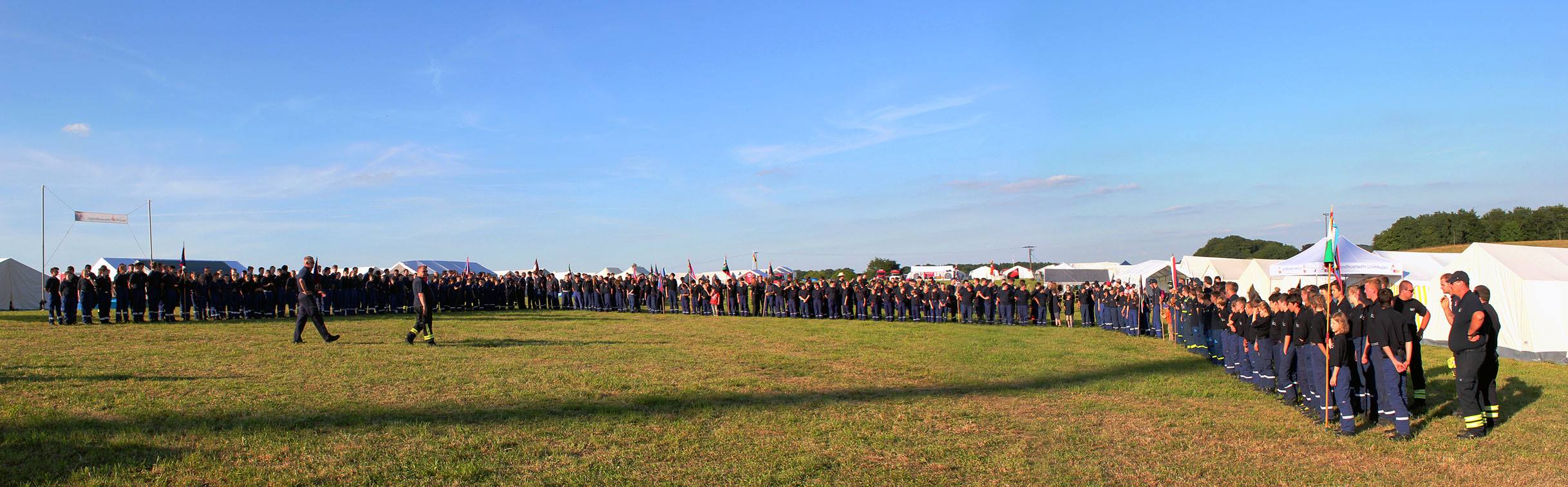 Eine große Gruppe von Menschen steht in mehreren Reihen auf einem offenen Feld, umgeben von Zelten. Der Himmel ist blau mit einigen Wolken, und die Teilnehmer scheinen an einem Event oder einer Versammlung teilzunehmen.